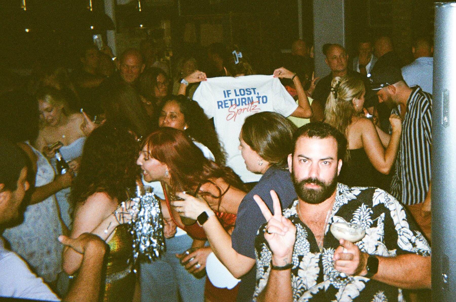 Crowded party with people mingling, a man in front making a peace sign holding a drink.